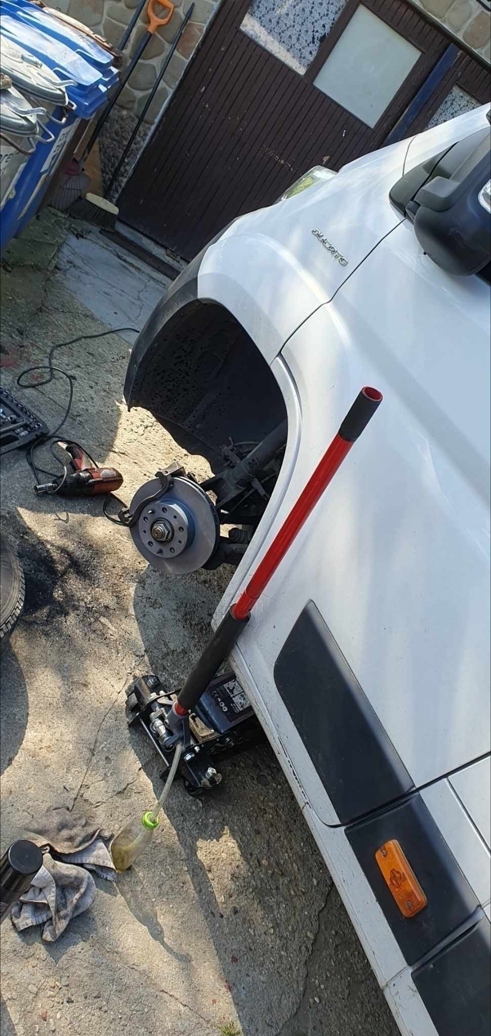 Biały samochód dostawczy na podnośniku hydraulicznym z odsłoniętą tarczą hamulcową, obok narzędzia warsztatowe i brudna szmata.