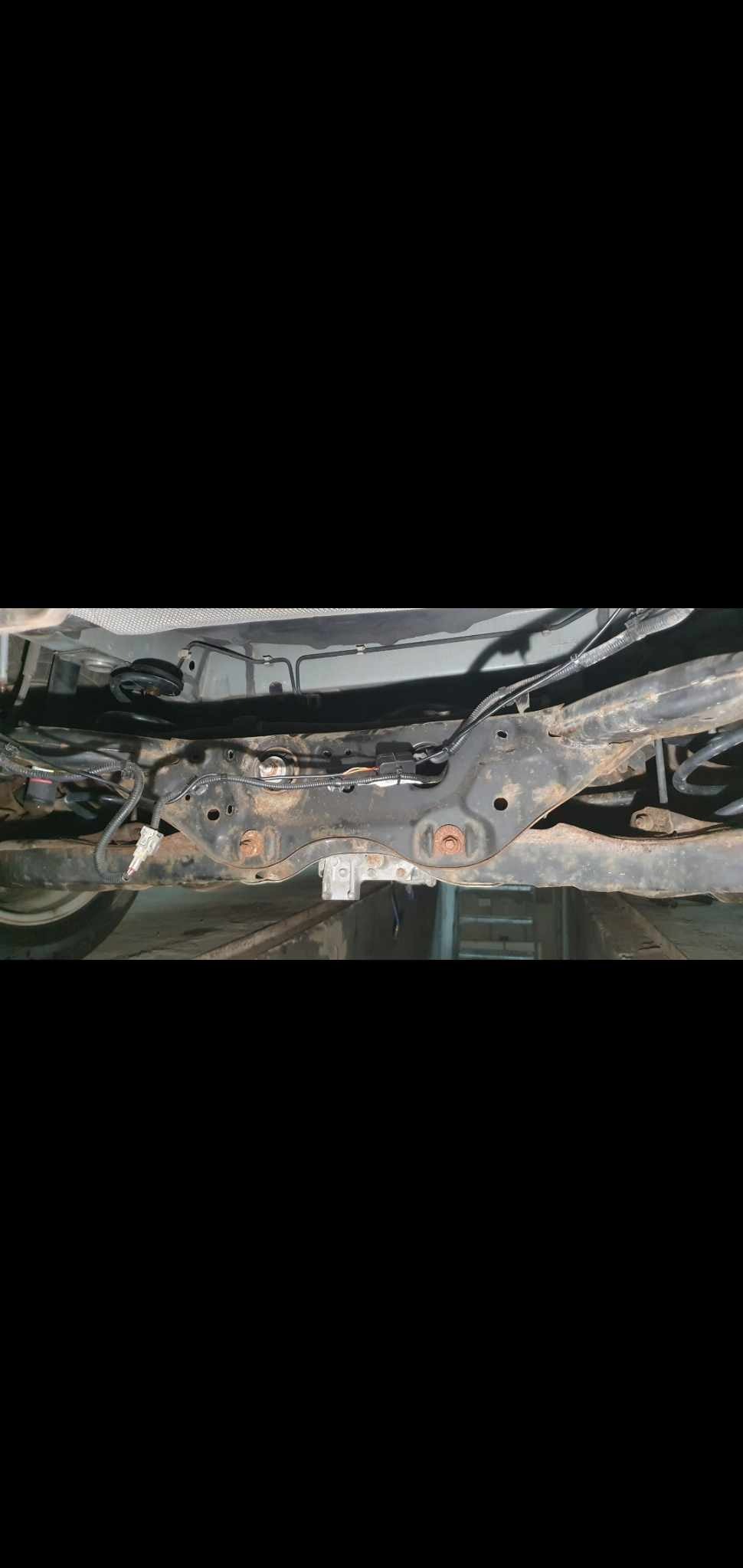 Widok podwozia samochodu z widocznymi elementami zawieszenia i przewodami, lekko zardzewiałe śruby mocujące.