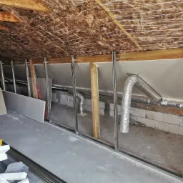 Poddasze w trakcie adaptacji: widoczne ocieplenie wełną mineralną pod dachem, metalowy szkielet ścian działowych, przewody wentylacyjne i płyty gipsowo-kartonowe czekające na montaż.