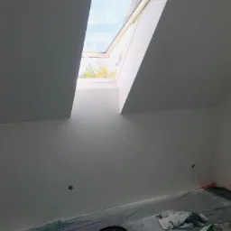 Poddasze z oknem dachowym po malowaniu na biało, zabezpieczona folią podłoga z widocznymi śladami farby, fragment nieotynkowanej ściany po prawej.