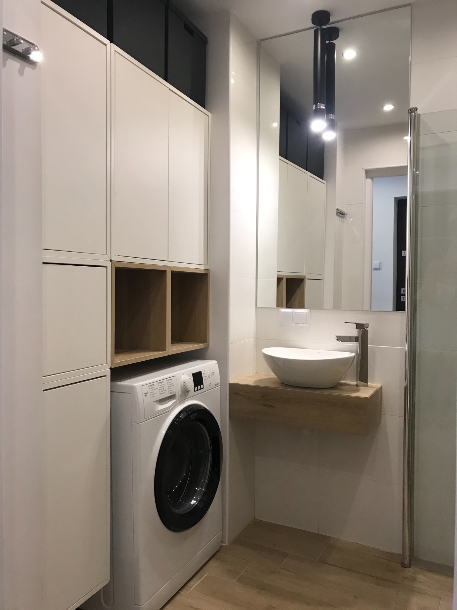 Nowoczesna łazienka z pralką Hotpoint, umywalką nablatową na drewnianym blacie i szafkami w zabudowie, oświetlenie punktowe.
