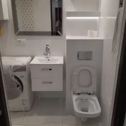 Nowoczesna łazienka z białą armaturą, umywalką z szafką, lustrem z podświetleniem, toaletą podwieszaną i pralką w folii ochronnej.