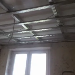 Metalowa konstrukcja podwieszanego sufitu w trakcie montażu, widoczne profile i częściowo zamontowane płyty, surowe ściany w tle.