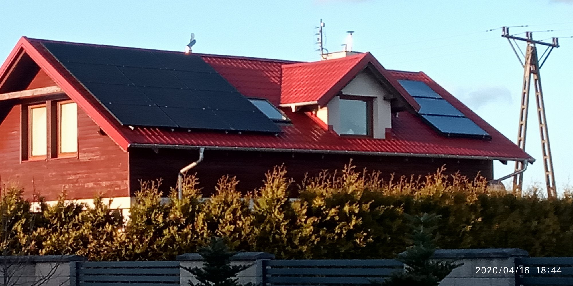 Czerwony dom jednorodzinny z panelami słonecznymi na dachu, widok zza żywopłotu, słup energetyczny w tle.