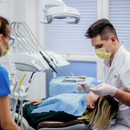 Dentysta w masce i rękawicach wykonuje zabieg na pacjentce leżącej na fotelu dentystycznym, asystuje mu pielęgniarka, w tle widoczne narzędzia dentystyczne i lampa.