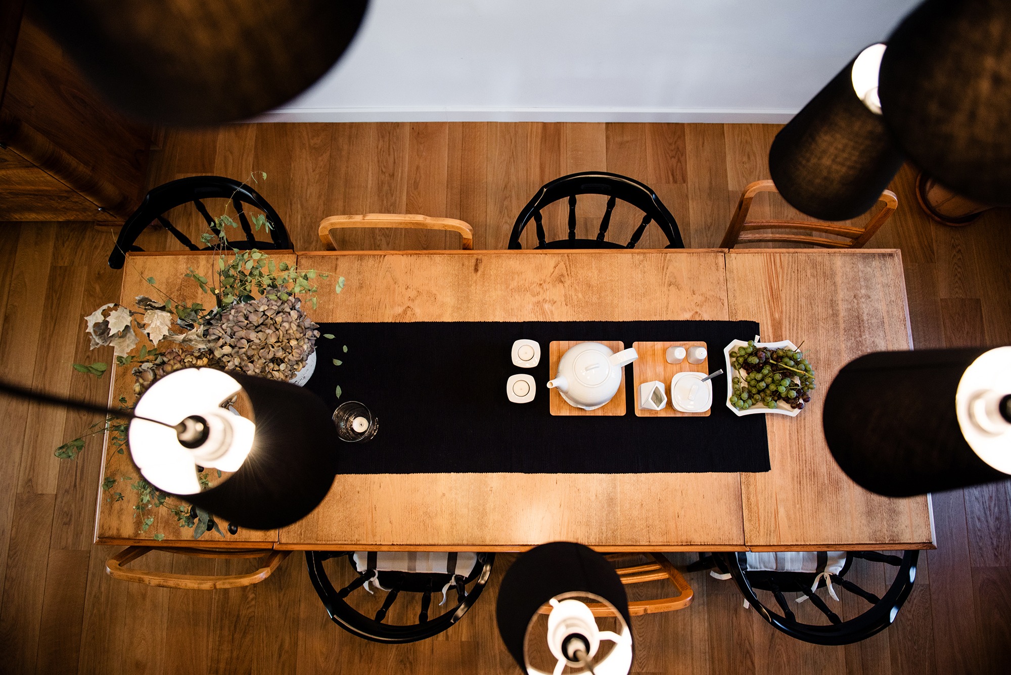 Widok z góry na drewniany stół z czarnym bieżnikiem, zastawą herbacianą, owocami i bukietem, otoczony krzesłami i lampami z czarnymi abażurami.