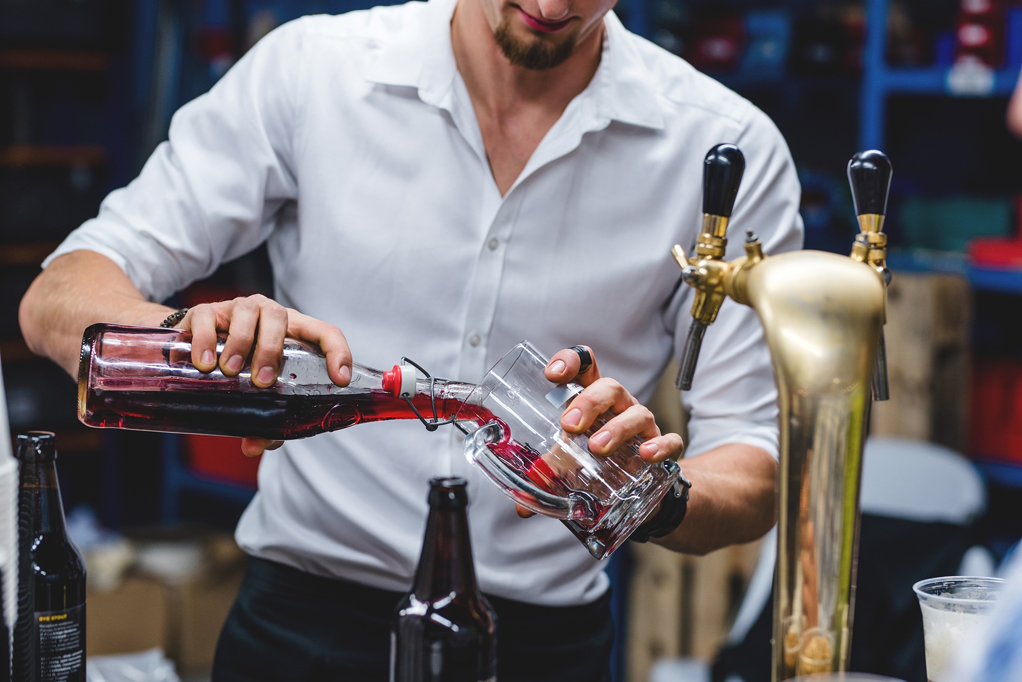 Barman w białej koszuli nalewa ciemnoczerwony napój z butelki typu swing-top do szklanki, obok widoczny kran do piwa i inne butelki, zdjęcie z eventu dla Wozijadło.pl.