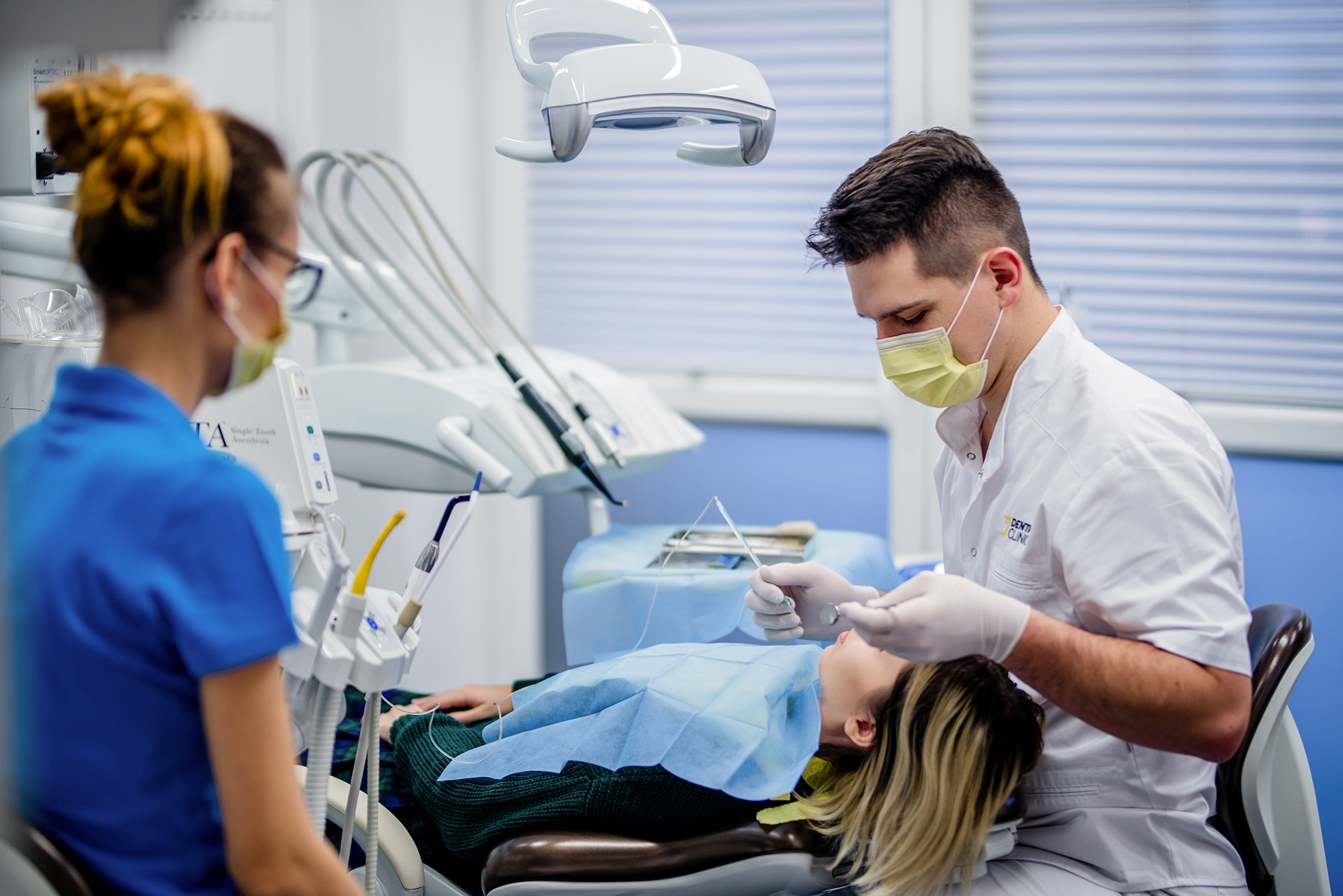 Dentysta w masce i rękawicach wykonuje zabieg na pacjentce leżącej na fotelu dentystycznym, asystuje mu pielęgniarka, w tle widoczne narzędzia dentystyczne i lampa.