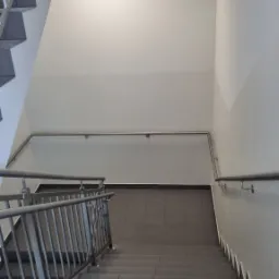 Klatka schodowa z szarymi schodami i metalową balustradą, pomalowane ściany na biało-szaro, widok z góry.