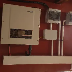 Zamontowany inwerter SOFAR Solar z podłączonymi przewodami i zabezpieczeniami na czerwonej ścianie, widoczny fragment osoby w kapturze.