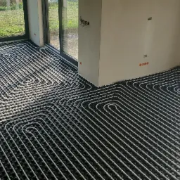 Białe rury ogrzewania podłogowego ułożone na czarnej macie izolacyjnej w nowym budynku, widoczne okna i przygotowane deski na zewnątrz.