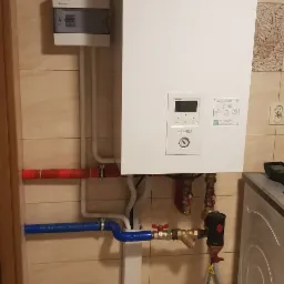 Biały piec gazowy Panasonic Aquarea Smart Cloud zamontowany na ścianie z beżowych płytek, widoczne rury w izolacji czerwonej i niebieskiej oraz skrzynka elektryczna obok.