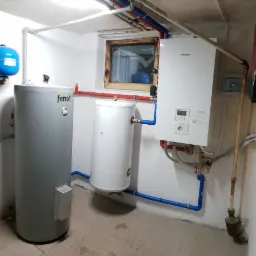 Instalacja grzewcza w piwnicy: szary zbiornik Ferolli, biały bojler i biały piec Panasonic Aquarea z rurami w kolorach czerwonym i niebieskim, widok na okno.