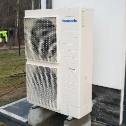 Zewnętrzna jednostka pompy ciepła Panasonic, zamontowana na betonowych podstawach przy budynku z elewacją wykończoną kamieniem dekoracyjnym, widoczne przewody i rura odprowadzająca skropliny.