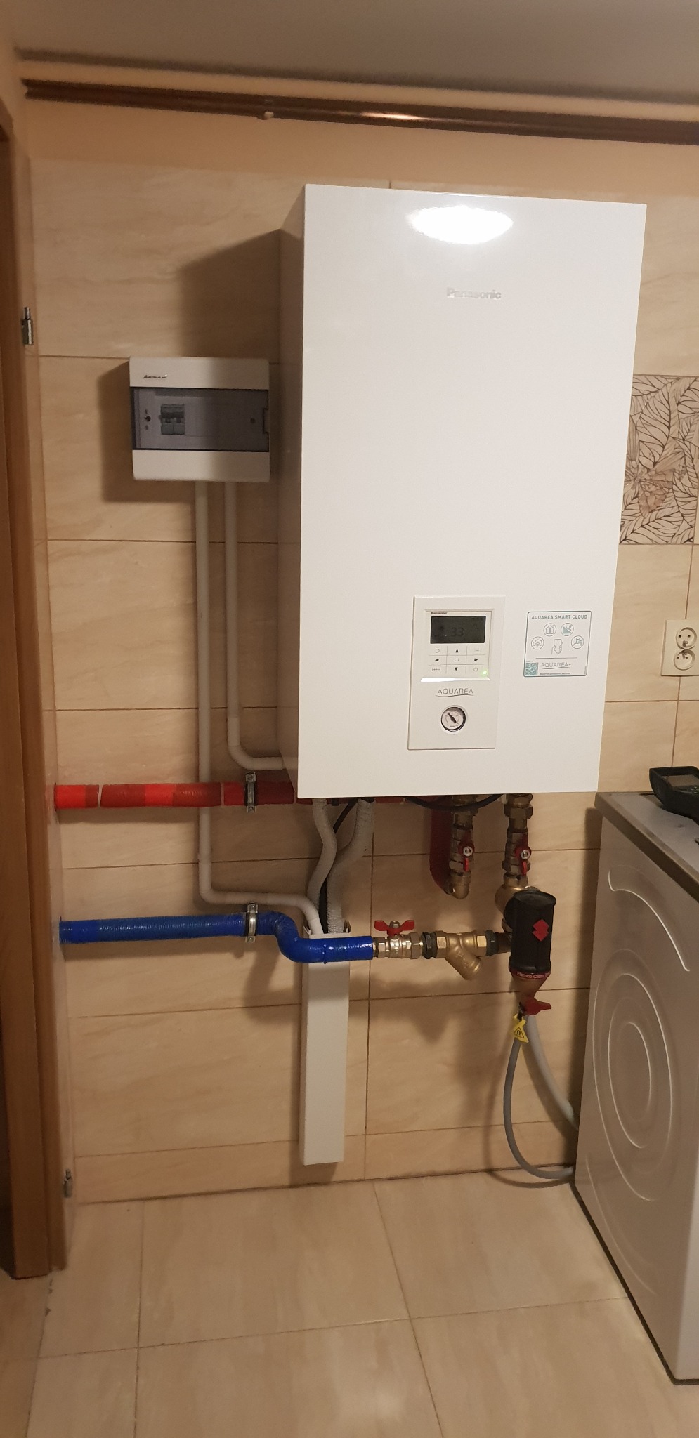 Biały piec gazowy Panasonic Aquarea Smart Cloud zamontowany na ścianie z beżowych płytek, widoczne rury w izolacji czerwonej i niebieskiej oraz skrzynka elektryczna obok.