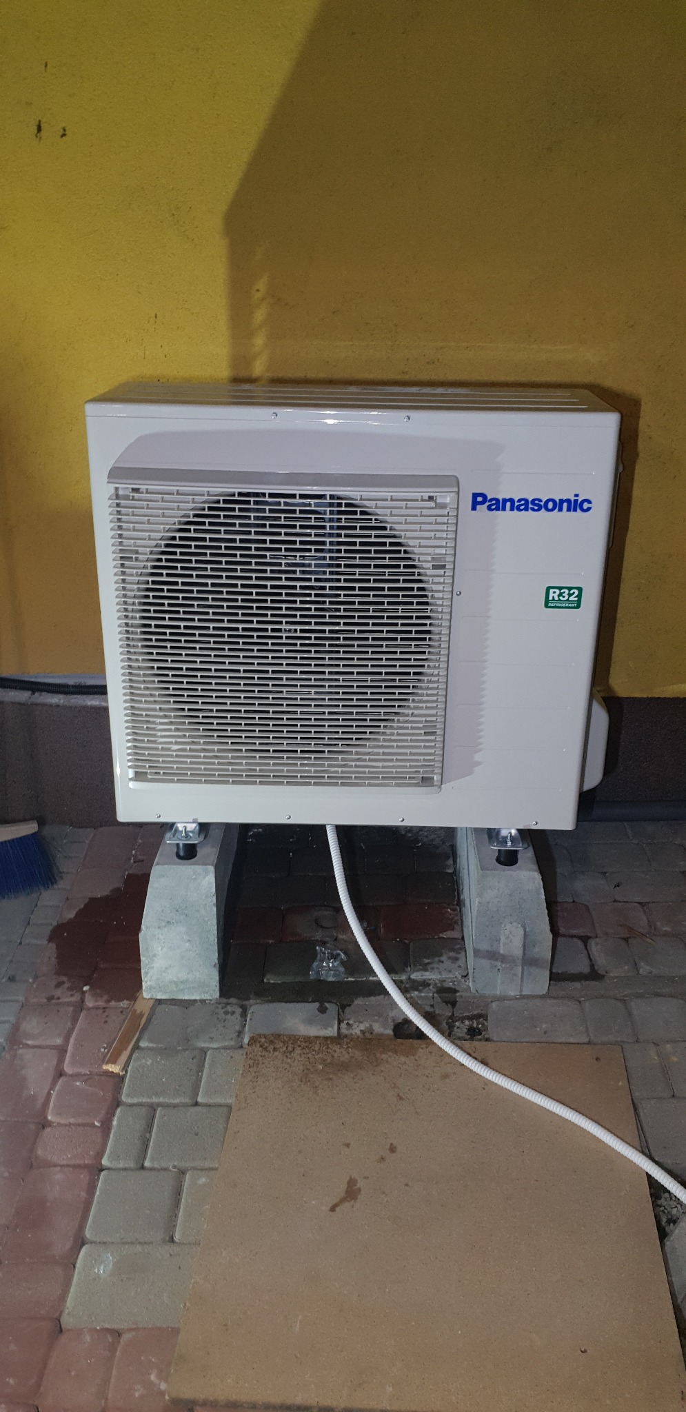 Zewnętrzna jednostka klimatyzacji Panasonic R32, umieszczona na betonowych podstawach, z widocznym przewodem chłodniczym, na tle żółtej ściany i kostki brukowej.