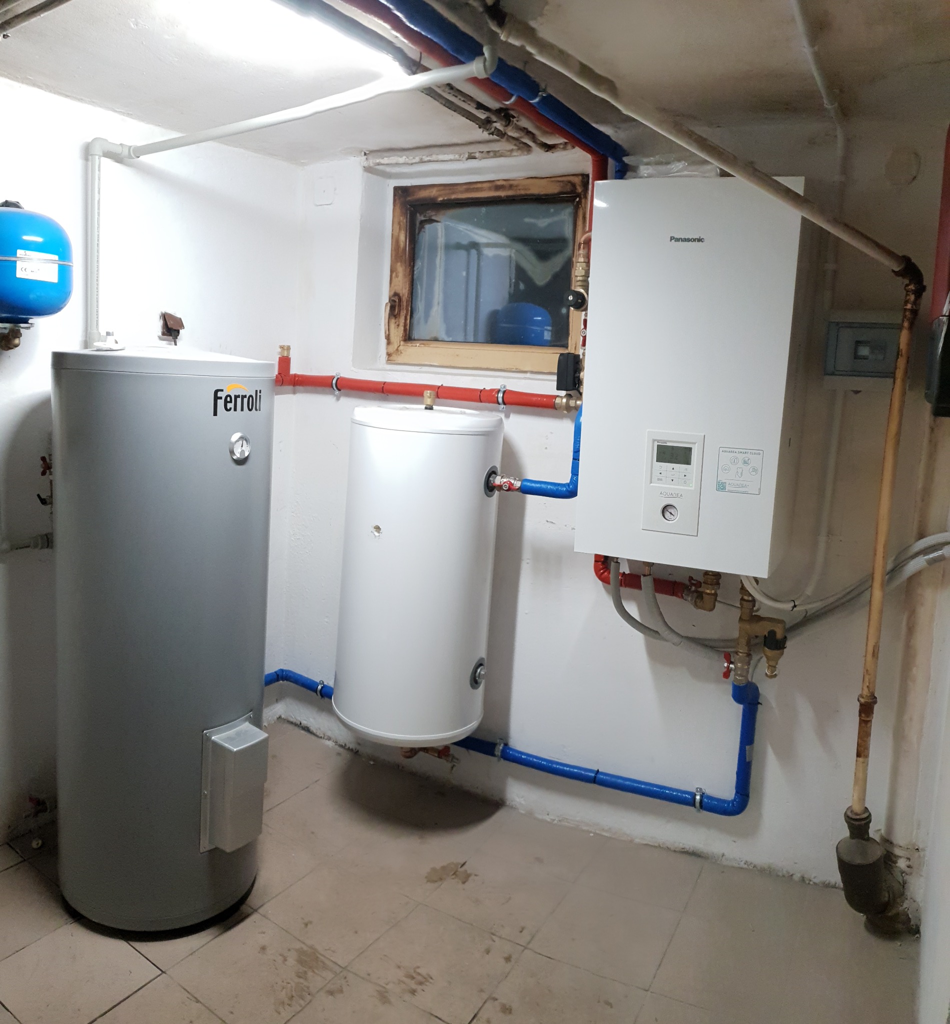 Instalacja grzewcza w piwnicy: szary zbiornik Ferolli, biały bojler i biały piec Panasonic Aquarea z rurami w kolorach czerwonym i niebieskim, widok na okno.