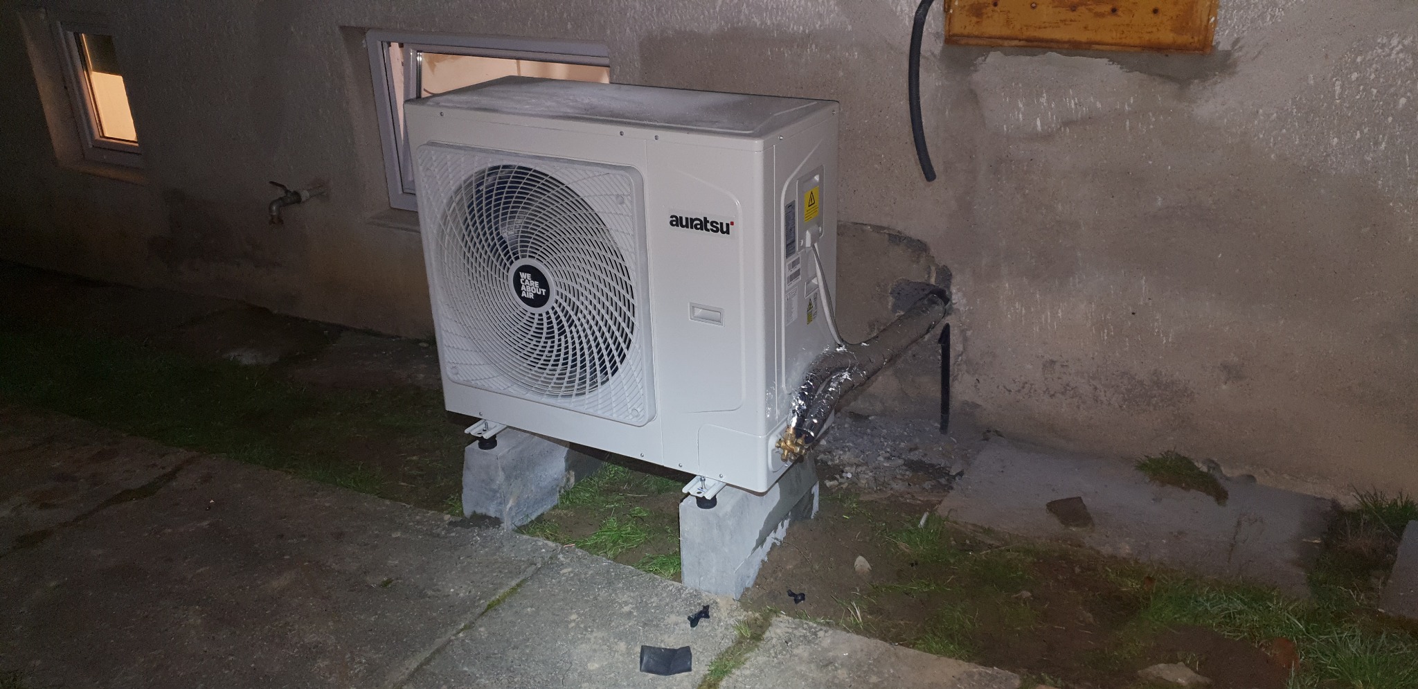 Zewnętrzna jednostka pompy ciepła marki Auratsu, zamontowana na betonowych bloczkach przy ścianie budynku z widocznymi oknami i rurami w izolacji.