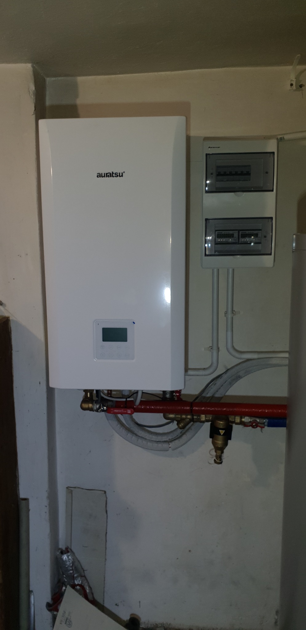 Biały, wiszący kocioł gazowy marki Auratsu z wyświetlaczem, podłączony do instalacji grzewczej z czerwonymi rurami, widoczne również szare, elastyczne przewody i skrzynka z bezpiecznikami na ścianie.