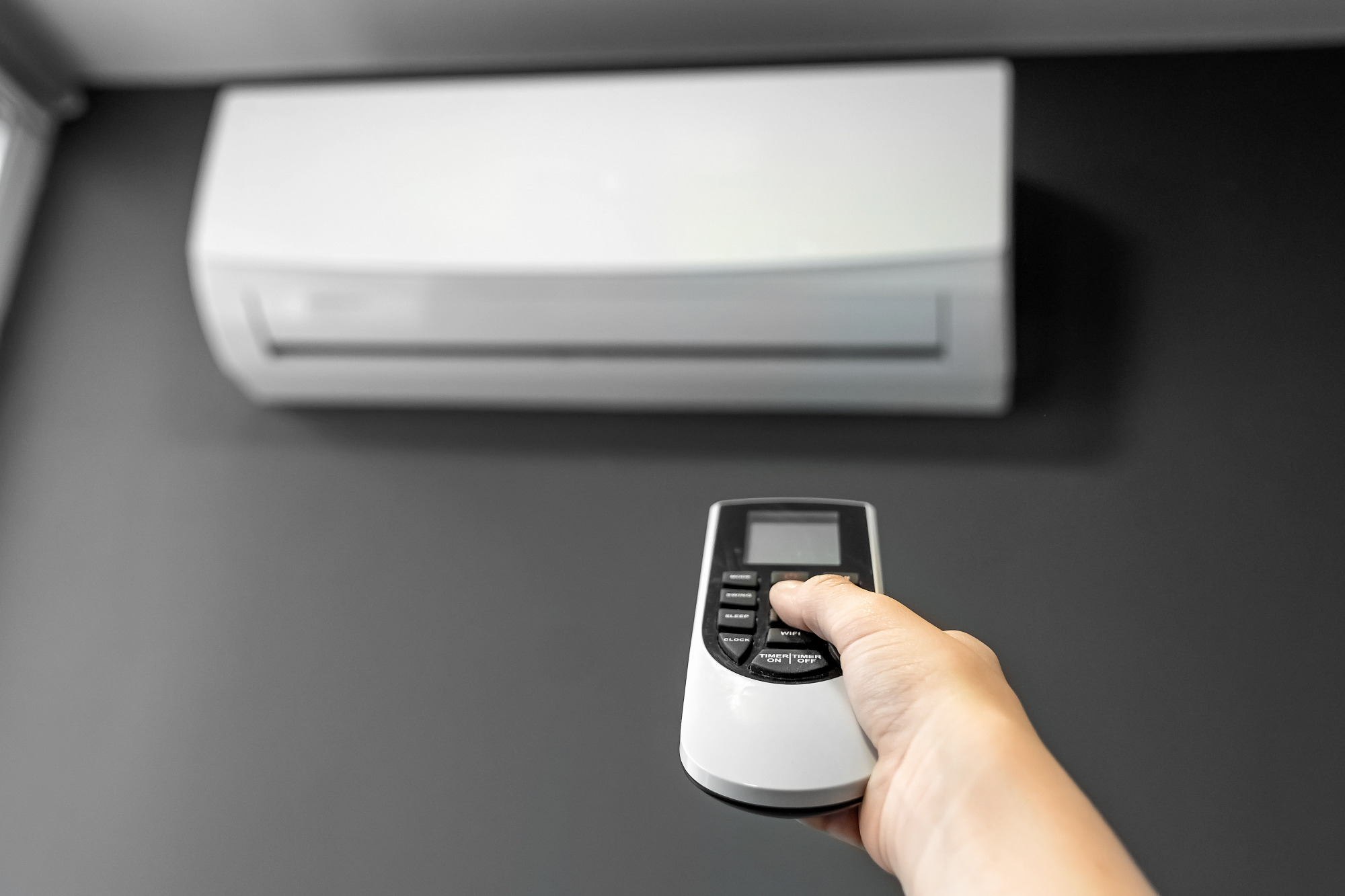 Biała klimatyzacja ścienna z pilotem w dłoni na tle szarej ściany.