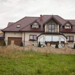 Dom jednorodzinny z beżową elewacją, brązowym dachem z dachówki, balkonem i dużym oknem panoramicznym, otoczony kamiennym ogrodzeniem z metalowymi przęsłami.