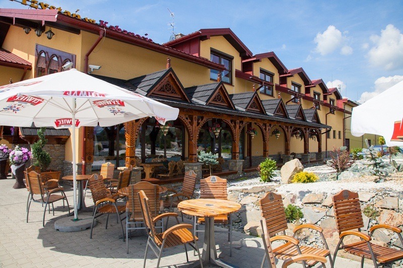 Elewacja budynku z drewnianą werandą, stolarka okienna widoczna w tle, drewniane meble ogrodowe na pierwszym planie, widok na zewnątrz z okien.