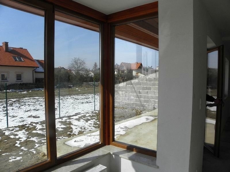 Duże przesuwne okna z brązowymi ramami w nowym domu, widok na ogród z resztkami śniegu i domy w tle.