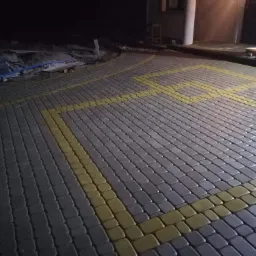 Ułożona kostka brukowa w odcieniach szarości z żółtymi liniami wyznaczającymi strefę; w tle widoczne materiały budowlane i fragment budynku.