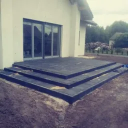 Nowoczesny taras z ciemnoszarej kostki brukowej z trzema schodami prowadzącymi do domu z dużymi przesuwnymi drzwiami, widoczne niedokończone prace ziemne wokół tarasu.