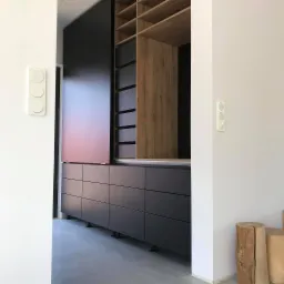 Częściowo otwarta, nowoczesna szafa wnękowa z przesuwnymi drzwiami, z czarnymi frontami szuflad i drewnianą wnęką z półkami, widziana z perspektywy korytarza z białymi ścianami i włącznikami światła.