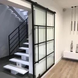 Nowoczesne schody z białymi stopniami i czarną balustradą, oddzielone od salonu przesuwnymi drzwiami z czarnymi szprosami i przezroczystym szkłem, widok z dołu.