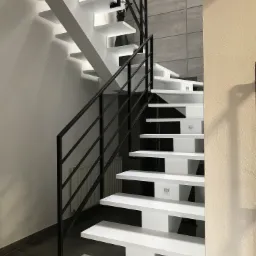 Nowoczesne schody wspornikowe z białymi stopniami i czarną metalową balustradą, oświetlenie punktowe pod stopniami, szara ściana w tle, logo firmy Chutzpa Design w prawym górnym rogu.