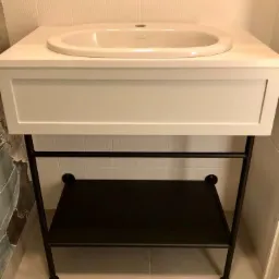 Biała umywalka nablatowa z szafką i czarnym metalowym stelażem z półką, na tle białych płytek.