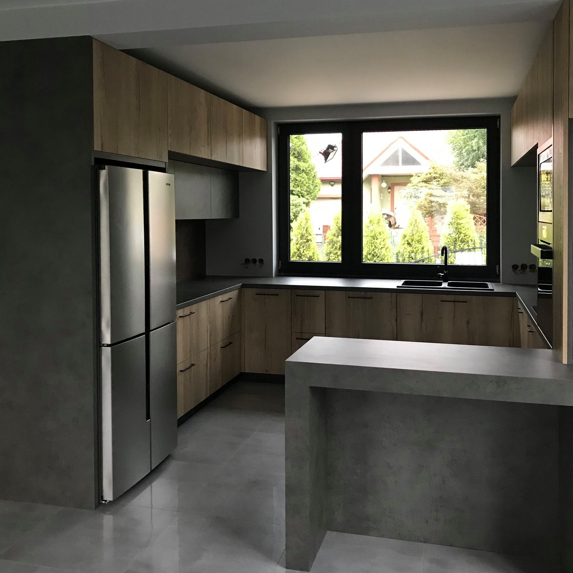 Nowoczesna kuchnia z drewnianymi frontami szafek, szarym blatem i wyspą, oraz dużą lodówką side-by-side, widok na ogród przez duże okno.