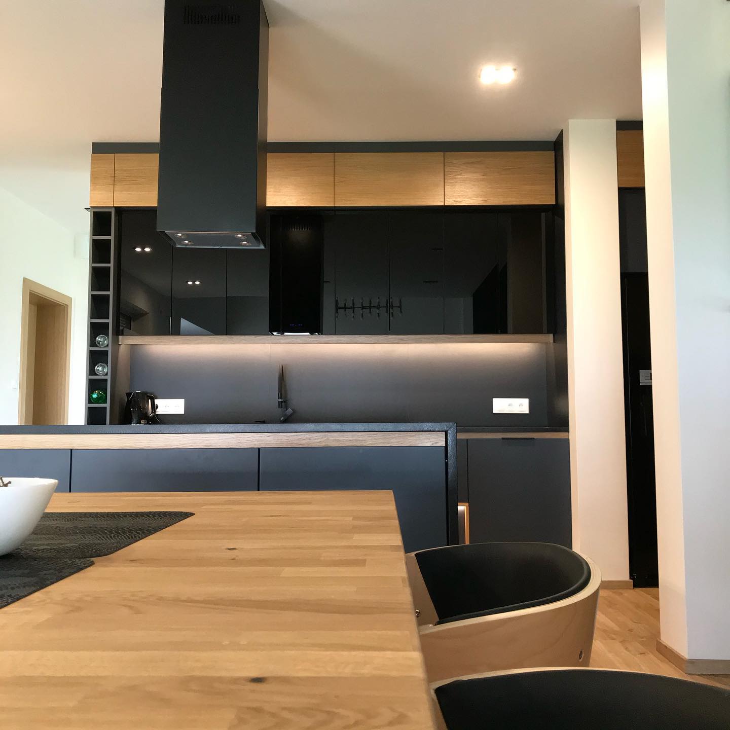 Nowoczesna kuchnia z czarnymi frontami szafek i drewnianymi akcentami, widok z perspektywy blatu stołu z drewnianym blatem, minimalistyczny design.