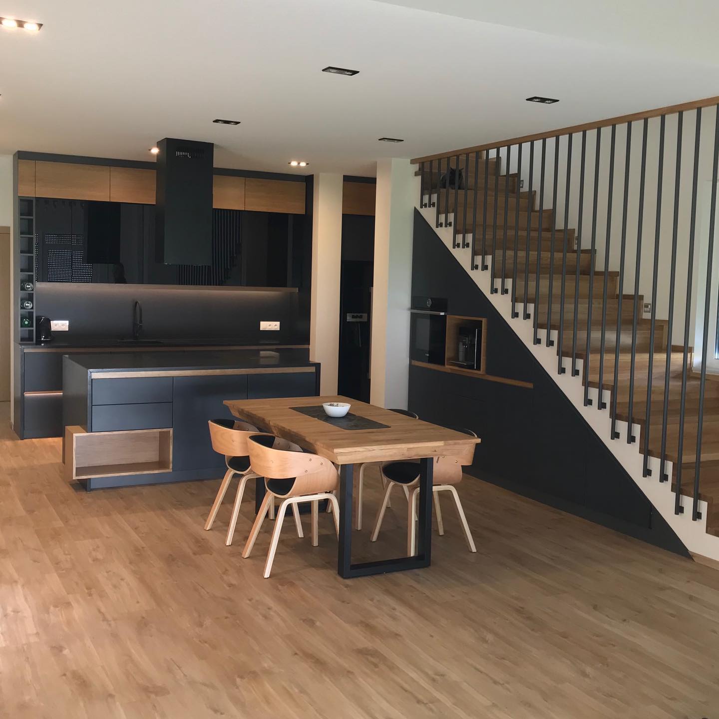 Nowoczesna kuchnia z wyspą, czarnymi frontami i drewnianymi akcentami, połączona z jadalnią ze stołem z blatem z surowego drewna i schodami z metalową balustradą, widok na czarnego kota na schodach.