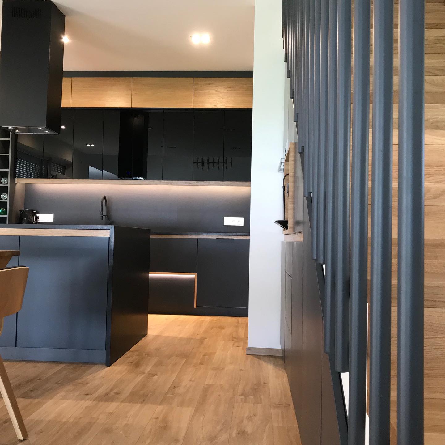 Nowoczesna, czarna zabudowa kuchenna z drewnianymi akcentami i podświetleniem LED, widok z perspektywy schodów z metalową balustradą.