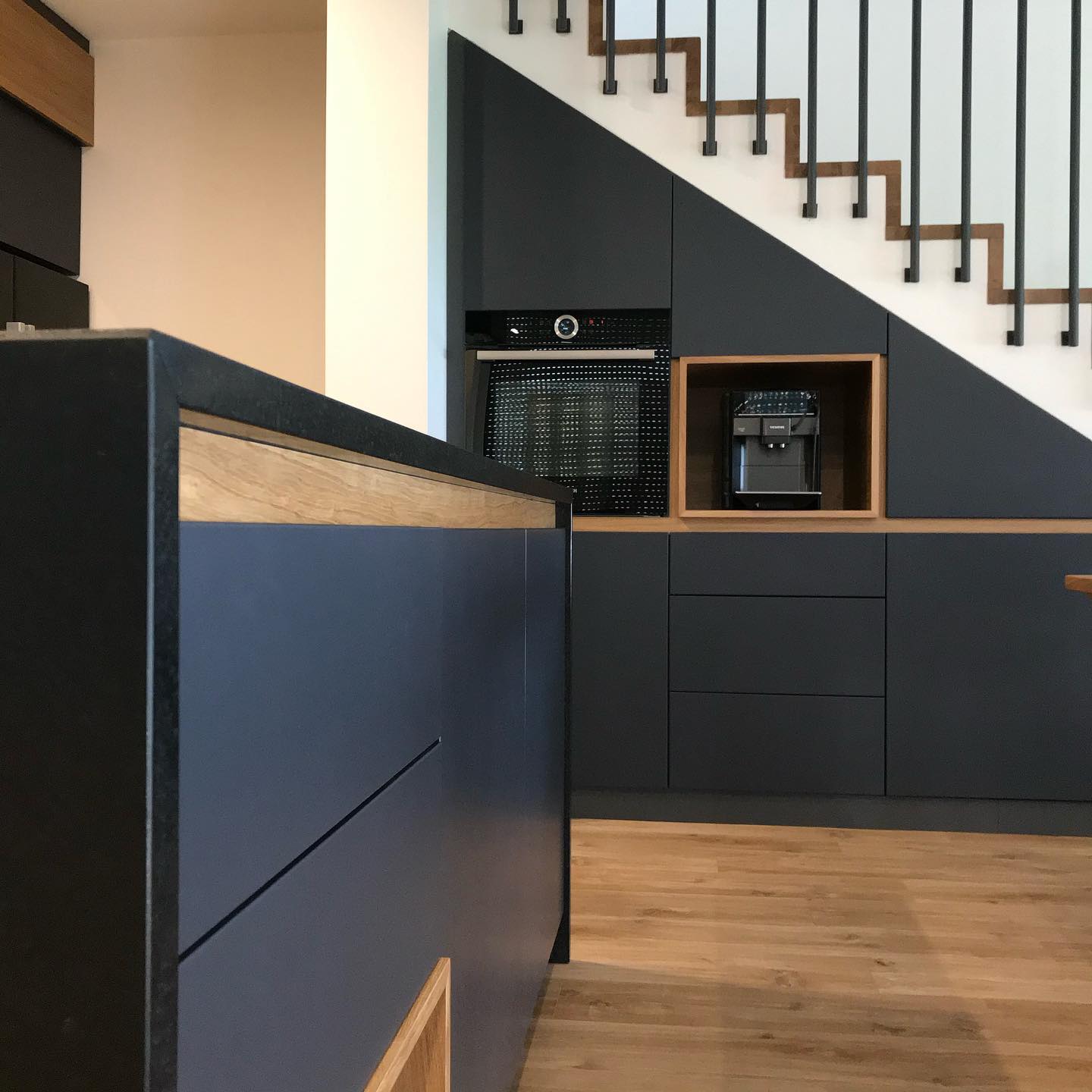 Ciemnogranatowa zabudowa kuchenna z drewnianymi akcentami, wkomponowana pod schody z czarnymi balustradami, z wbudowanym piekarnikiem i ekspresem do kawy.