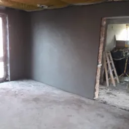 Wnętrze pokoju w stanie surowym po nałożeniu gładzi gipsowej, widoczne ślady zaprawy na podłodze, żółta izolacja na suficie i ceglana ościeżnica drzwiowa