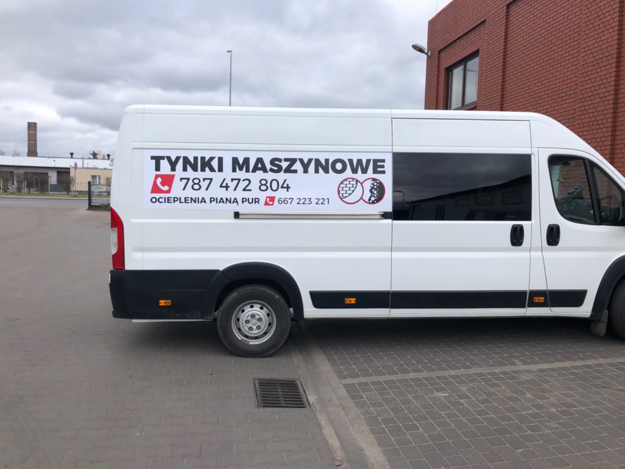 Biały samochód dostawczy z reklamą usług tynkarskich i ocieplania pianą PUR, zaparkowany na kostce brukowej, z budynkiem z czerwonej cegły w tle.