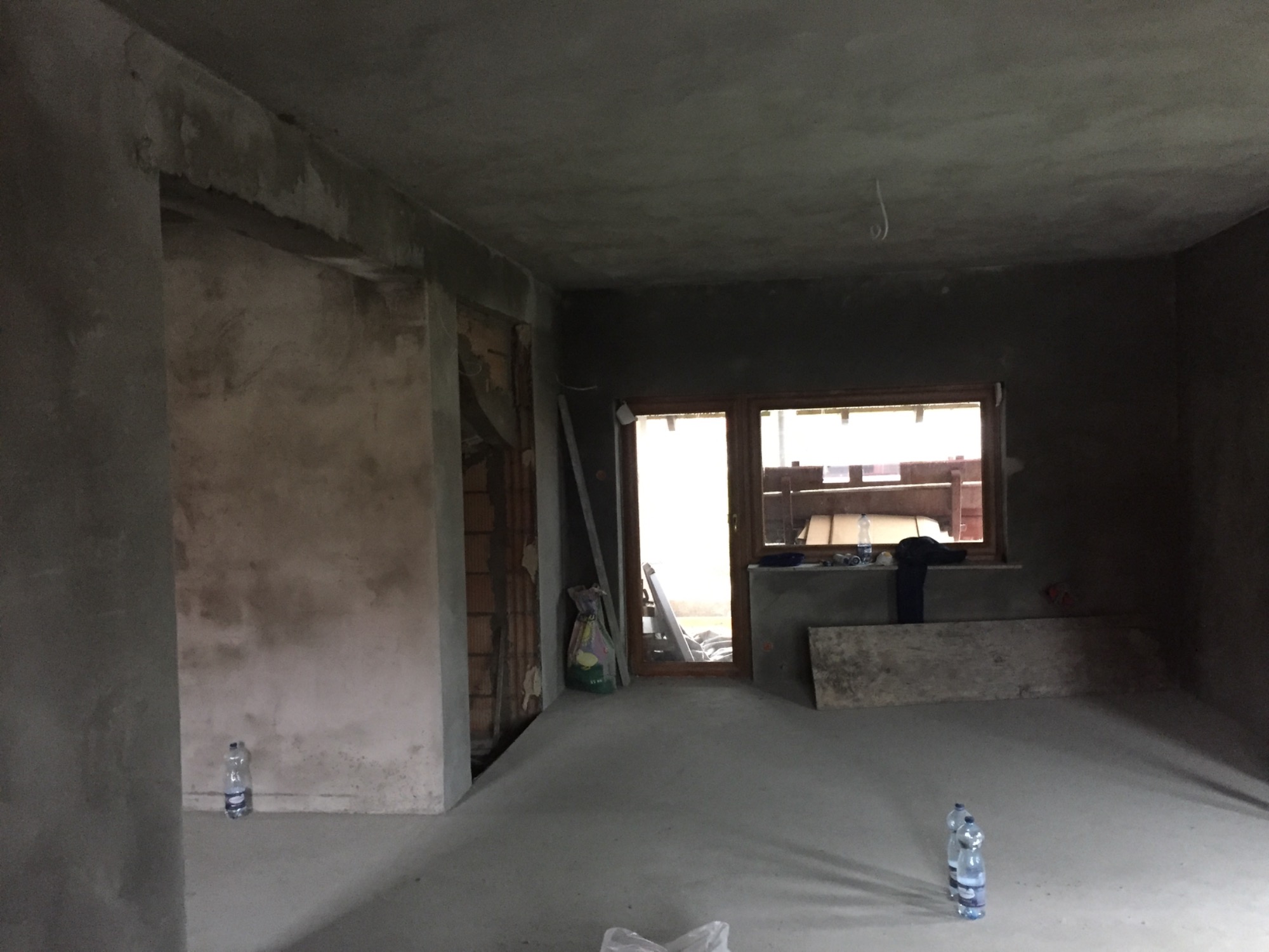 Surowe wnętrze po nałożeniu tynków: widoczne nierówności, butelki z wodą na podłodze, okno z widokiem na zewnątrz, ściany w szarym odcieniu, przygotowane do dalszych prac wykończeniowych.