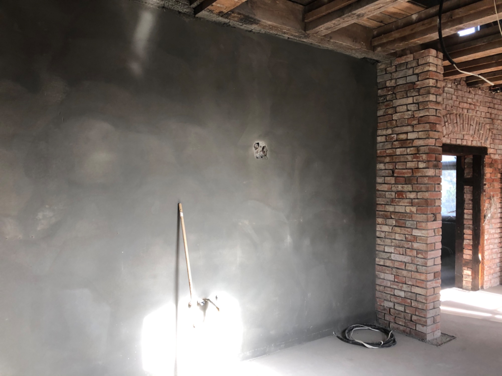 Świeżo nałożony, szary tynk maszynowy na ścianie z widocznym otworem na gniazdko elektryczne, z narzędziem do tynkowania opartym o ścianę i fragmentem ceglanej kolumny w tle.