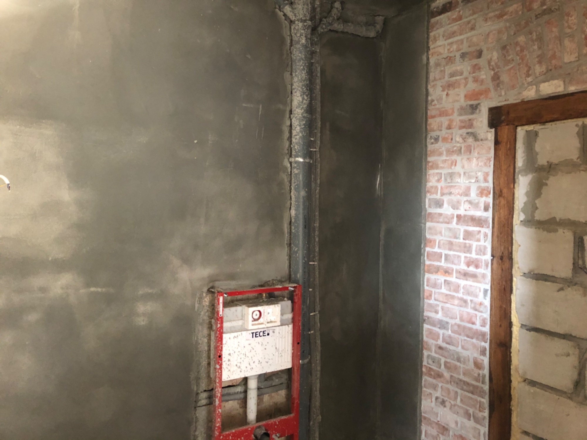 Surowa ściana po nałożeniu szarej zaprawy, widoczna instalacja sanitarna w czerwonej ramie, pionowa rura i fragment ściany z czerwonej cegły z drewnianą framugą.