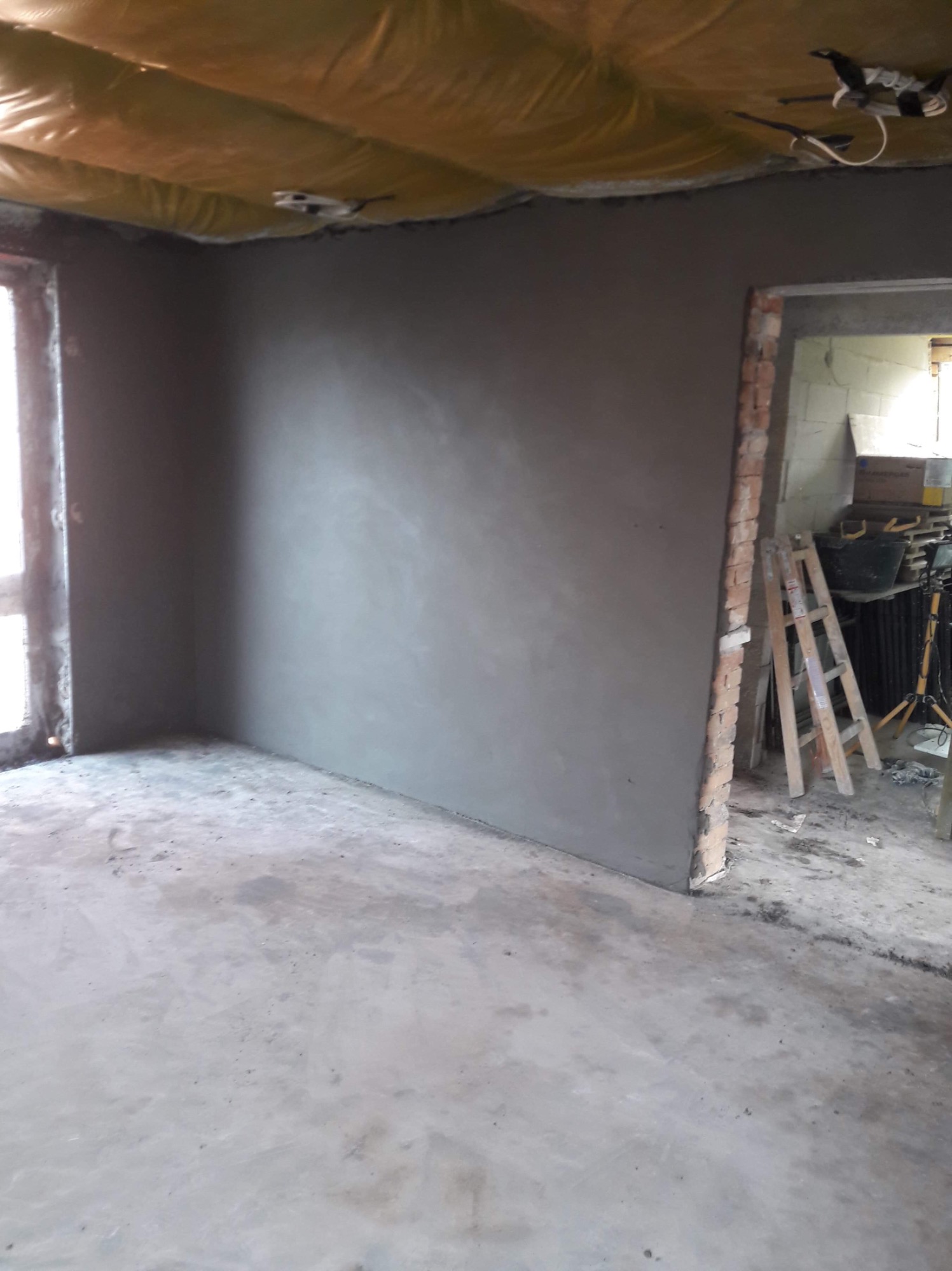 Wnętrze pokoju w stanie surowym po nałożeniu gładzi gipsowej, widoczne ślady zaprawy na podłodze, żółta izolacja na suficie i ceglana ościeżnica drzwiowa