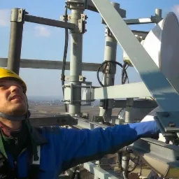 Instalator w kasku i uprzęży, na wysokości, montuje antenę telekomunikacyjną na metalowej konstrukcji, widok na miasto w tle.