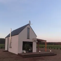 Niewielki, nowoczesny dom z antresolą, z białą elewacją, grafitowym dachem, kominem ze stali nierdzewnej, anteną satelitarną na dachu i drewnianą pergolą, oświetlony lampkami gruntowymi, z rowerem...