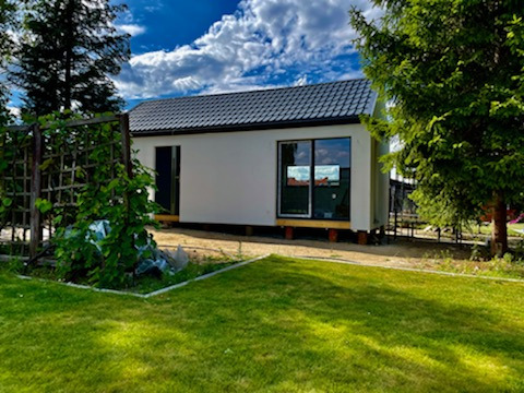 Nowoczesny, parterowy budynek z płaskim dachem, posadowiony na drewnianych legarach, widoczny z perspektywy ogrodu w słoneczny dzień.