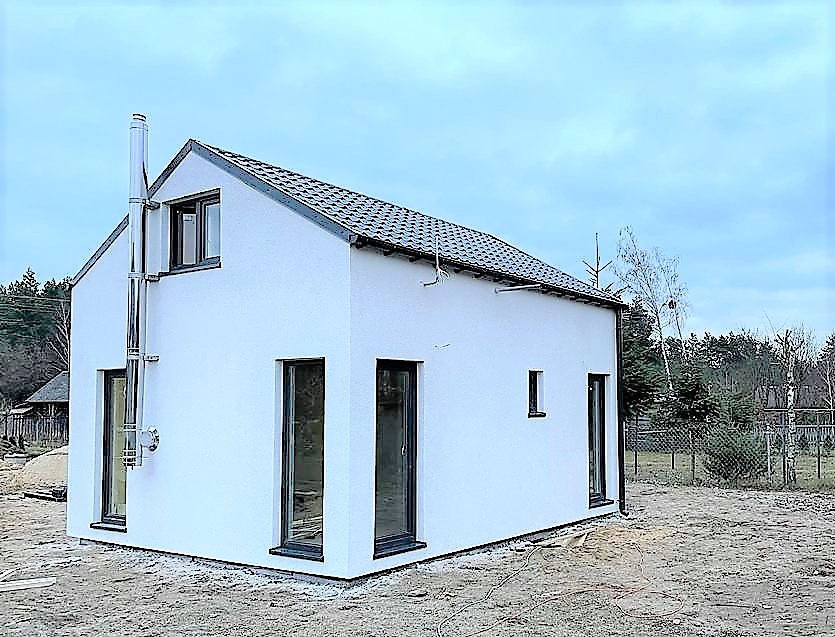 Niewielki, biały budynek z dwuspadowym dachem pokrytym szarą dachówką, z wystającym kominem ze stali nierdzewnej, widok z zewnątrz na tle lasu.