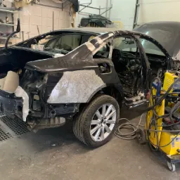 Czarny samochód w trakcie naprawy karoserii w warsztacie, z otwartą maską i bagażnikiem, z widocznymi elementami szpachli i spawarką GYS na wózku.