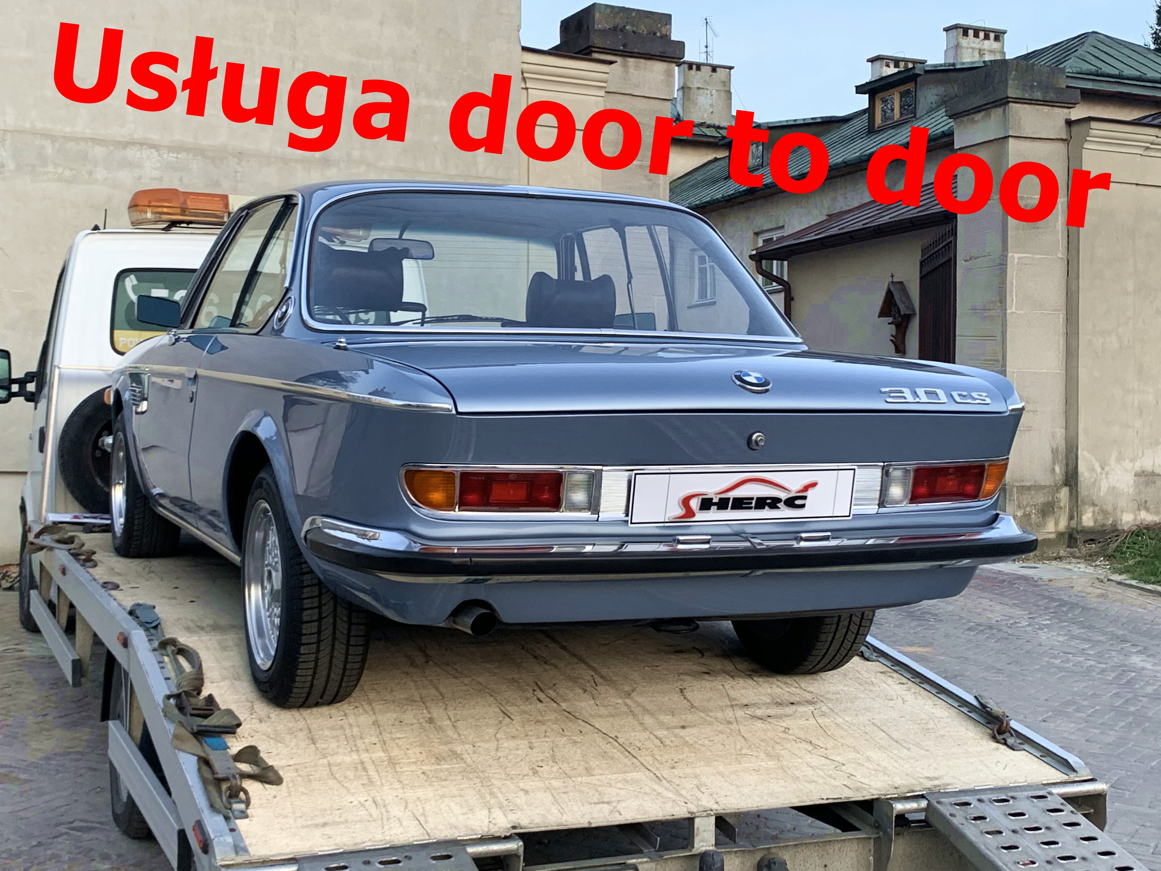 Błękitny samochód BMW 3.0 CS z lat 70. na platformie lawety, widok z tyłu, z logo firmy transportowej na tablicy rejestracyjnej.